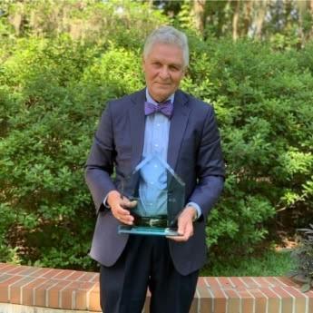 Dr. William “Bill” Mendenhall in an outdoor setting wearing a suit and bowtie on the occasion of receiving in 2020 a lifetime achievement award, which he holds in his hands.